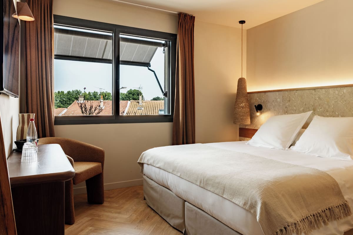 A hotel room with a bed, a desk, a chair, a window with curtains, and a lamp.