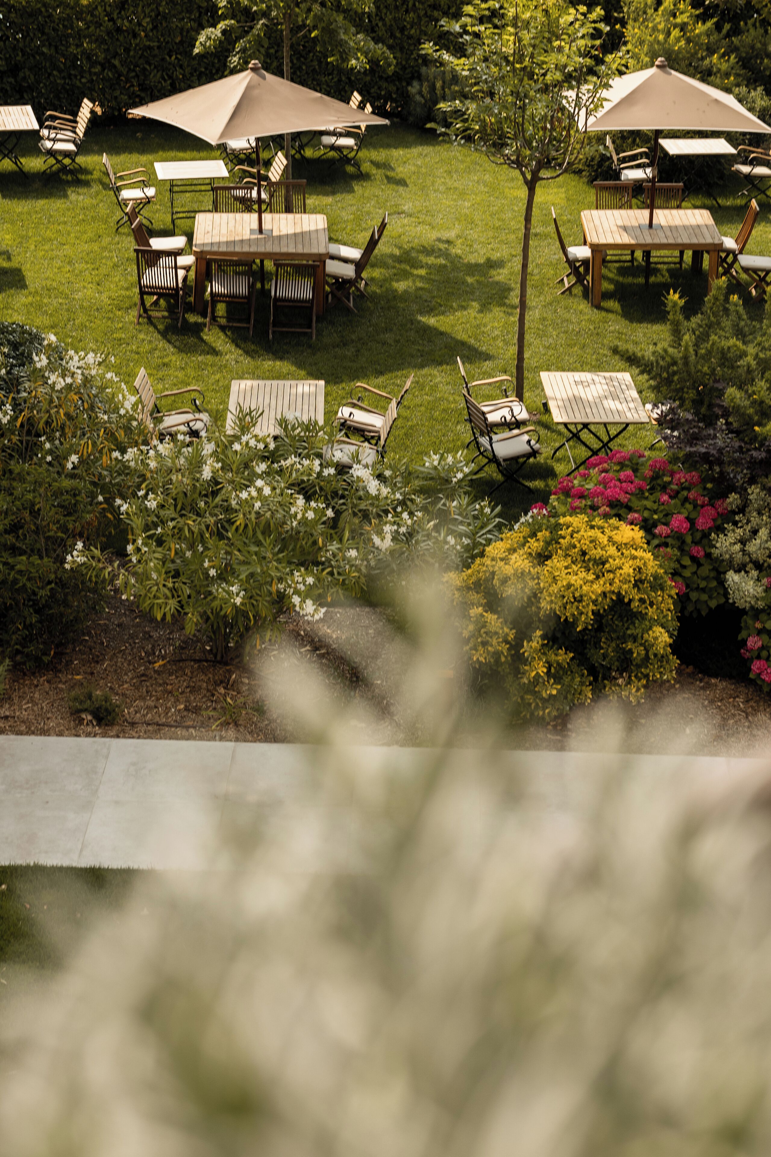 An outdoor patio area with multiple wooden tables and chairs, surrounded by lush greenery and flowering plants.