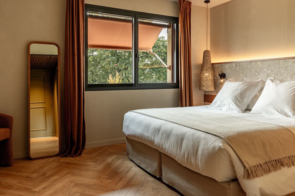A bedroom with a bed, a window with curtains, a mirror, and a wall-mounted light fixture.