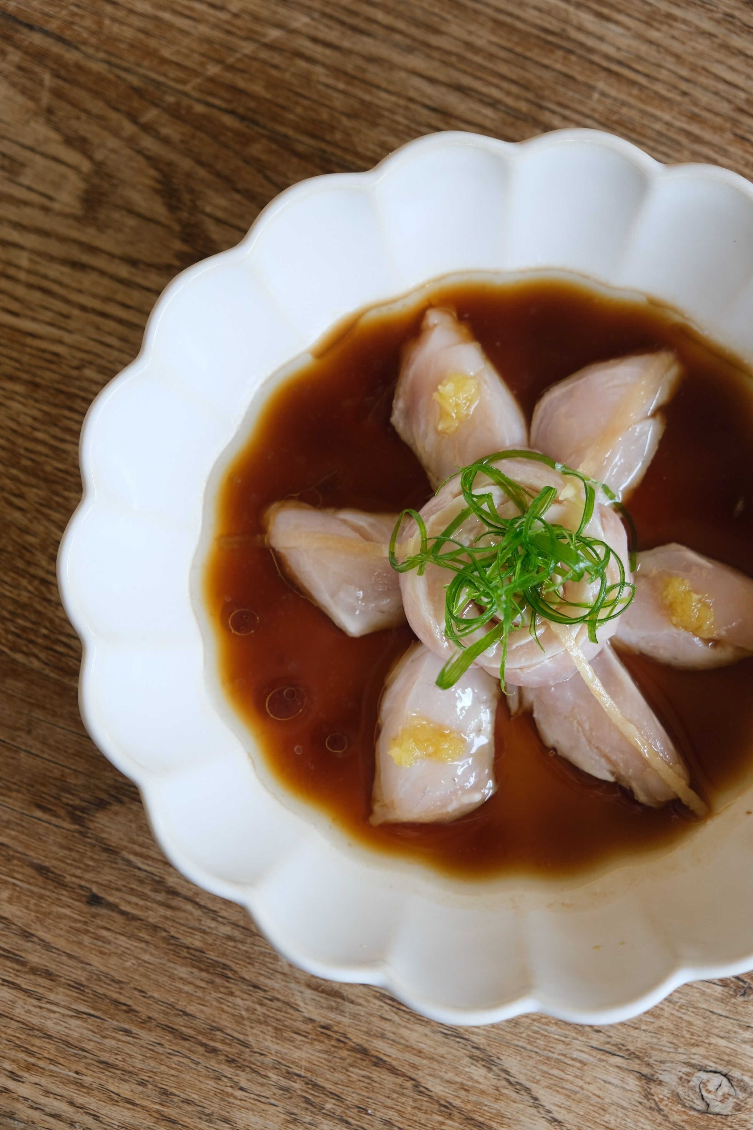 A white bowl with a scalloped edge containing sliced fish and green garnish in a brown sauce, placed on a wooden surface.