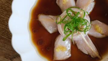 A white bowl with a scalloped edge containing sliced fish and green garnish in a brown sauce, placed on a wooden surface.