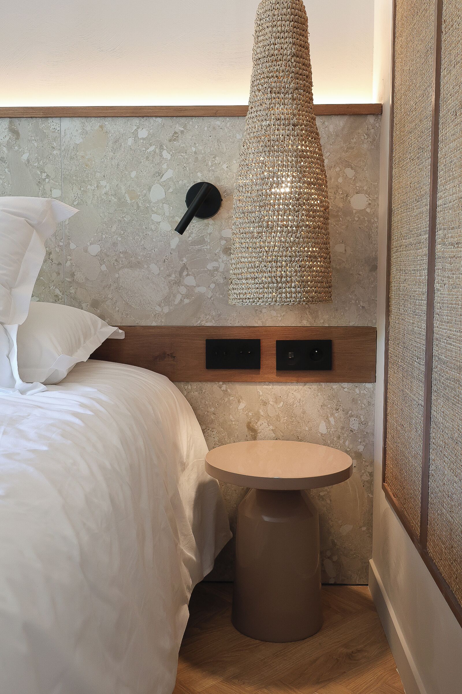 A bedroom with a bed, a round table, a textured wall hanging, and a wooden headboard.