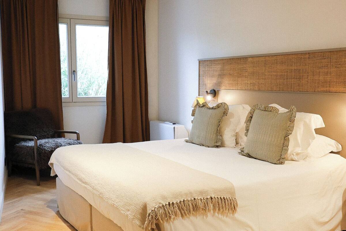 A bedroom with a bed, a chair, a window with curtains, and a bedside table.