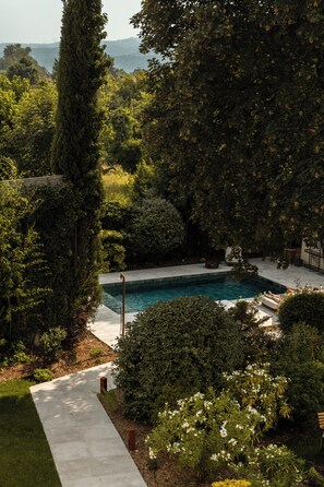 A rectangular swimming pool surrounded by lush greenery, with a concrete pathway leading to it.