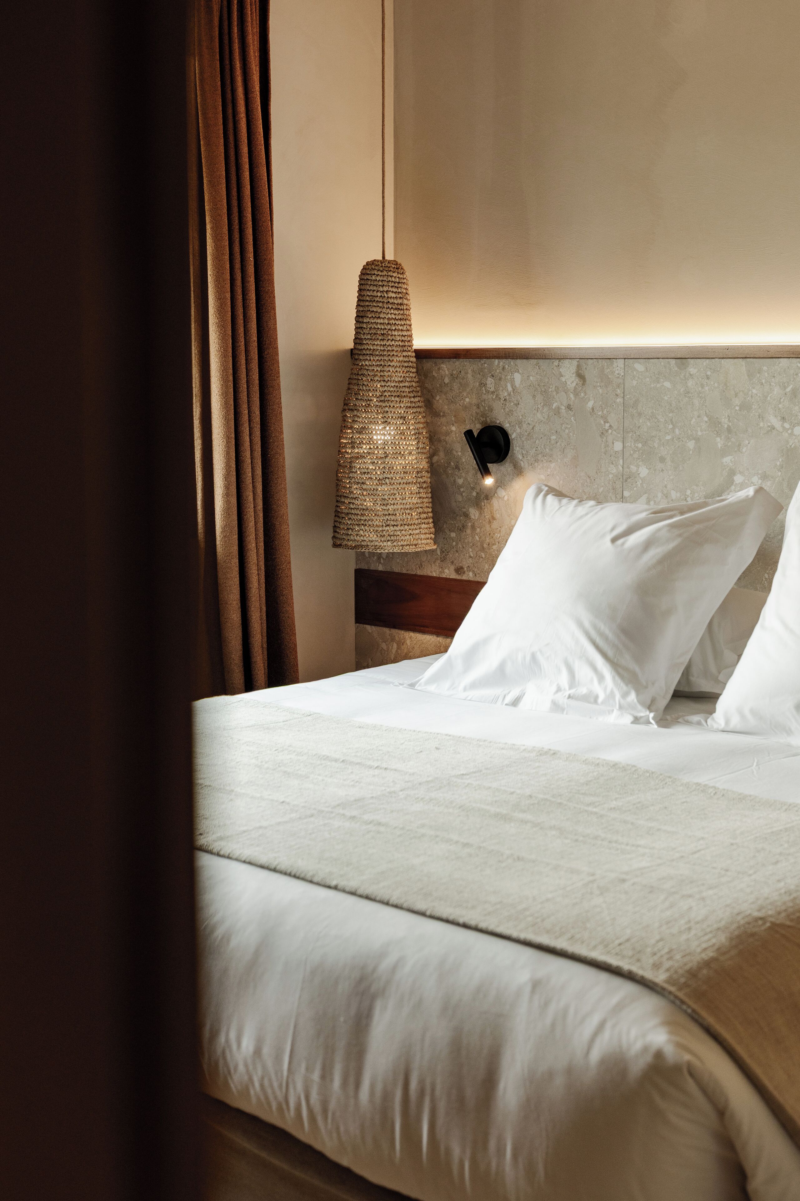A neatly made bed with white linens, a textured wall lamp, and a dark curtain partially drawn.