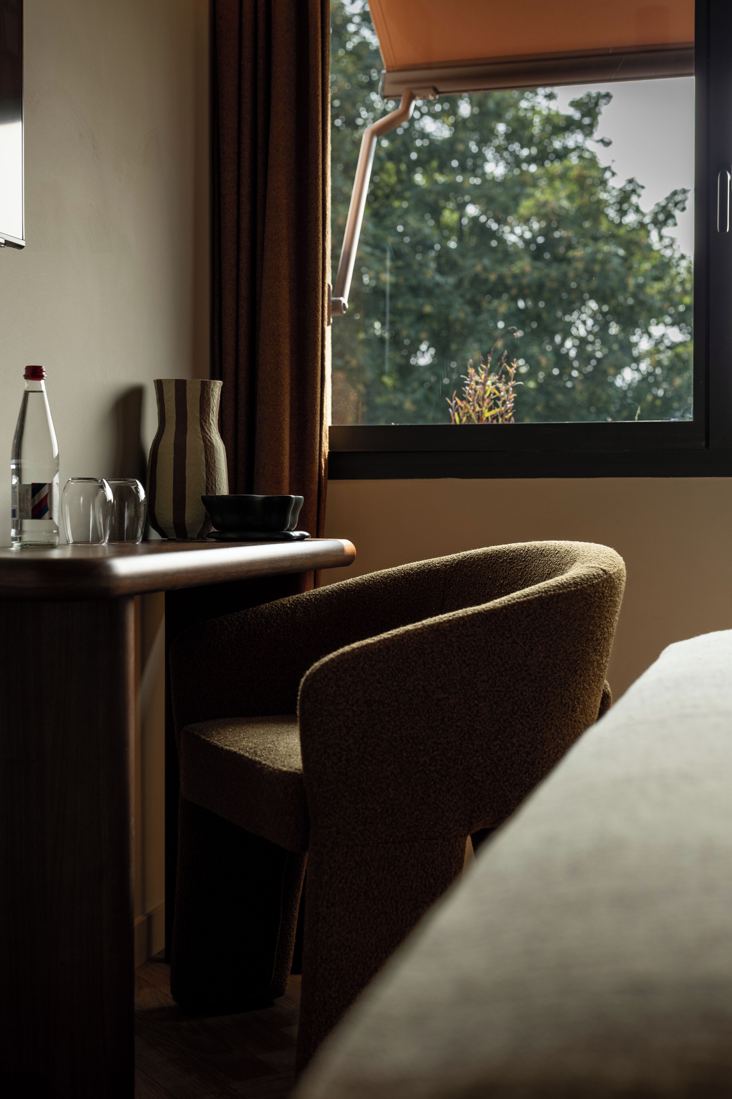 A hotel room with a bed, a desk with a chair, a window with a view of trees, and a curtain.
