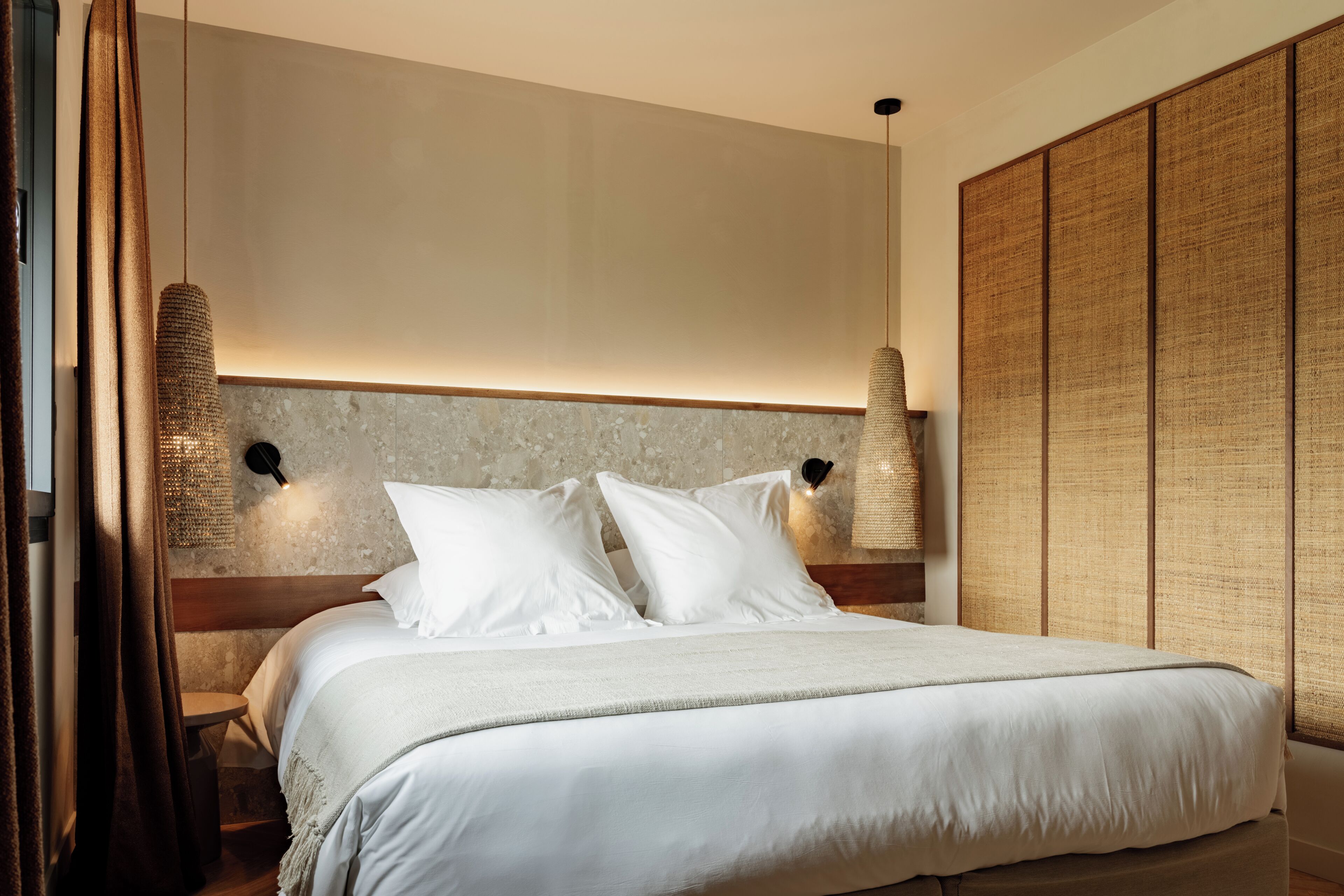 A neatly made bed with white linens, two wall-mounted lamps, and a large woven partition.
