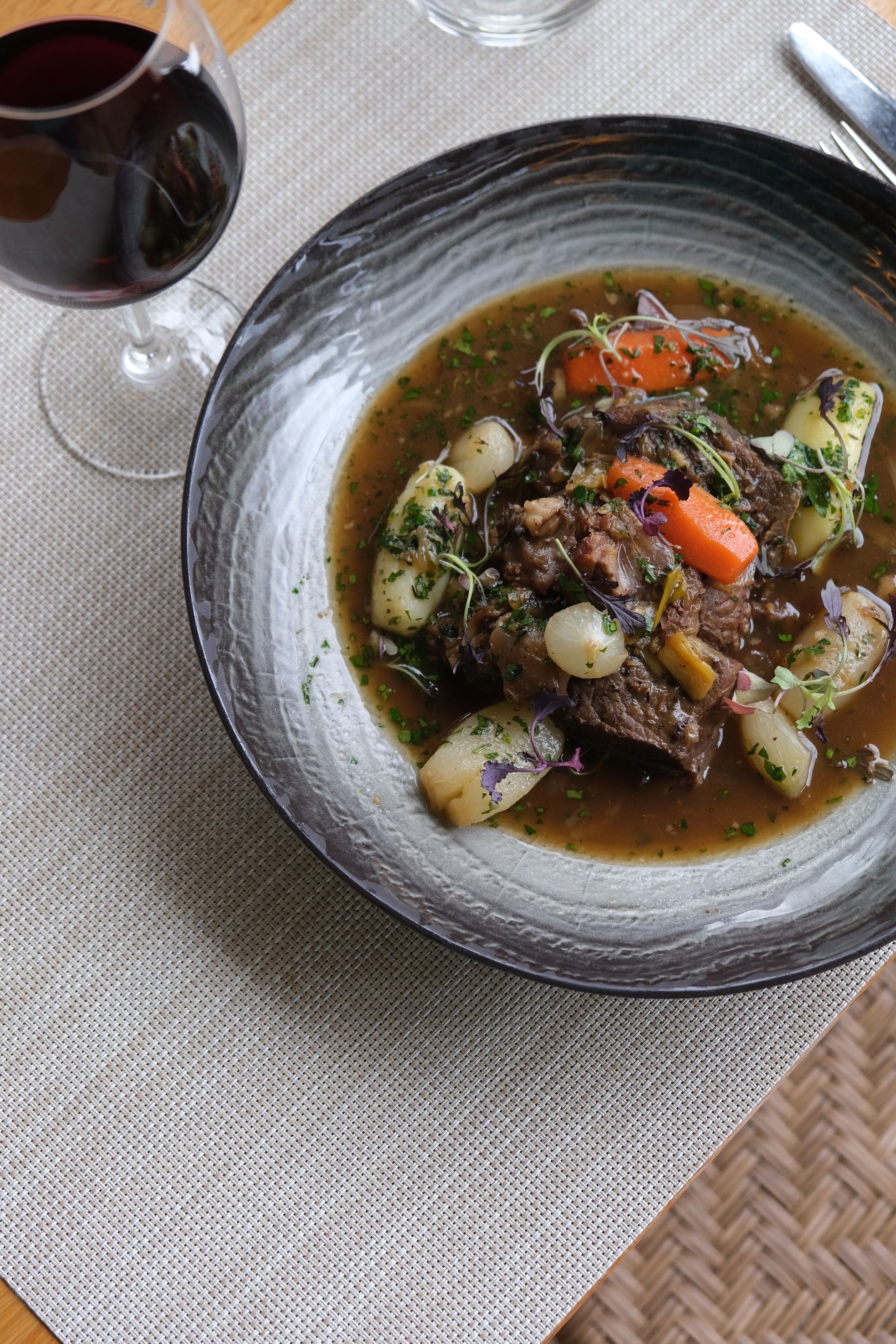 A bowl of beef stew with vegetables and a glass of red wine on a textured tablecloth.