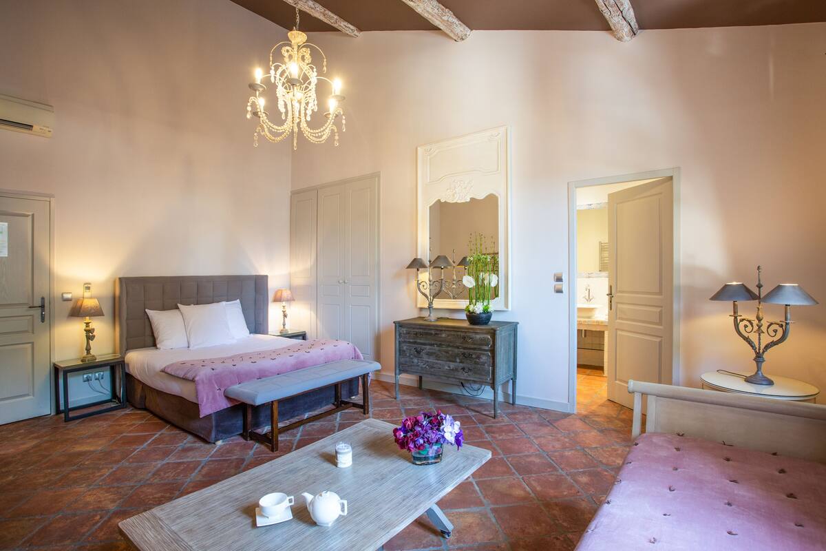A bedroom with a bed, bedside tables, a dresser, a bench, a table with a vase of flowers, and a chandelier.