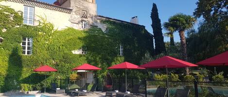 Una piscina con sillas de descanso y sombrillas rojas, rodeada de abundante vegetación y un edificio histórico con una torre.
