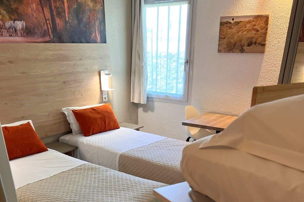 A hotel room with two beds, a small table, a chair, and a window with curtains.