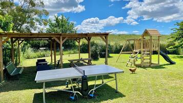 A covered outdoor area with a ping pong table, picnic tables, and a wooden play structure.