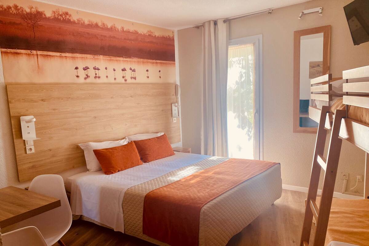 A hotel room with a wooden headboard, a bed with orange bedding, a desk with a chair, and a large window with curtains.