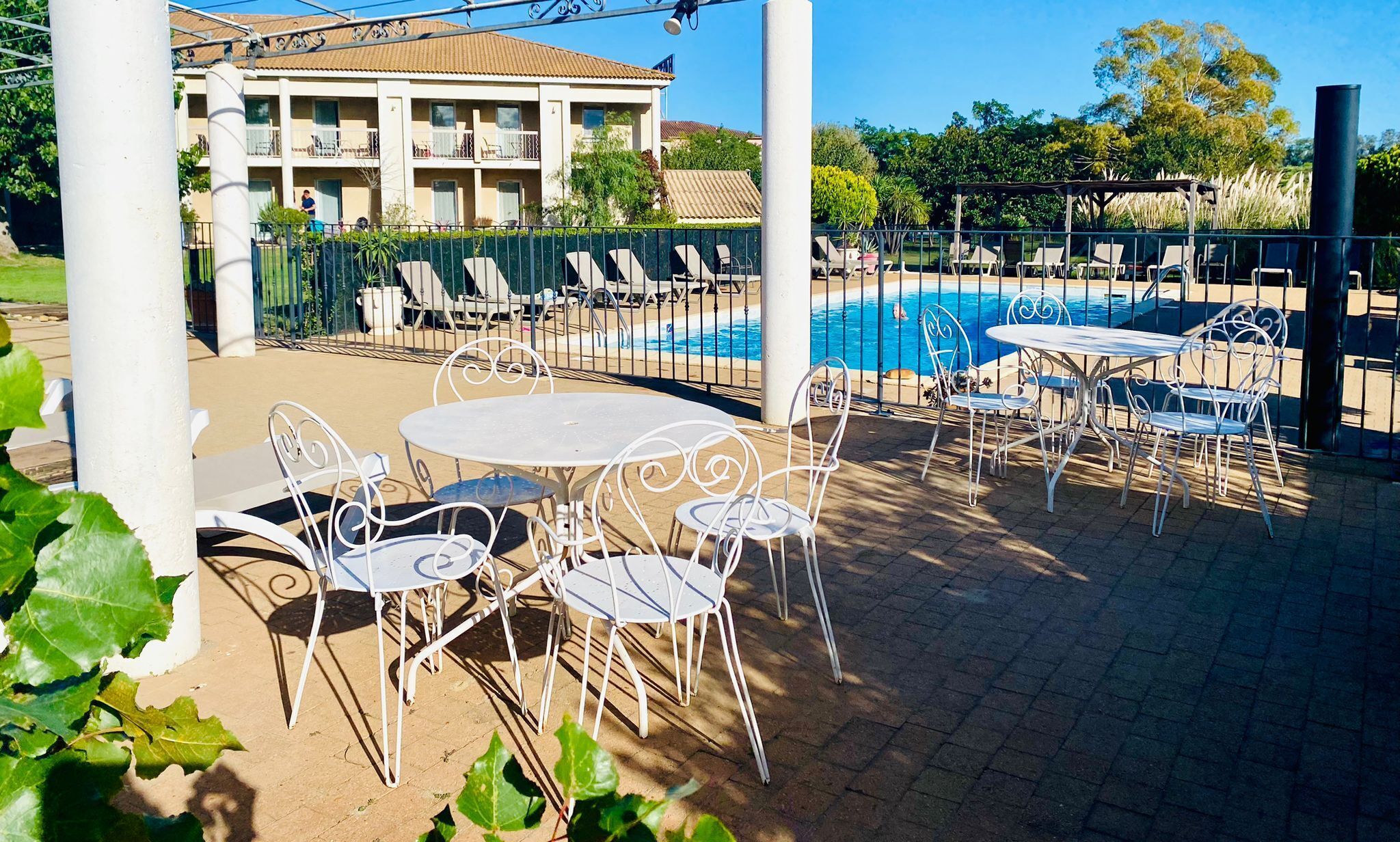 Un espace aménagé au bord de la piscine, avec des chaises et des tables en métal blanc, une piscine et un bâtiment en arrière-plan.