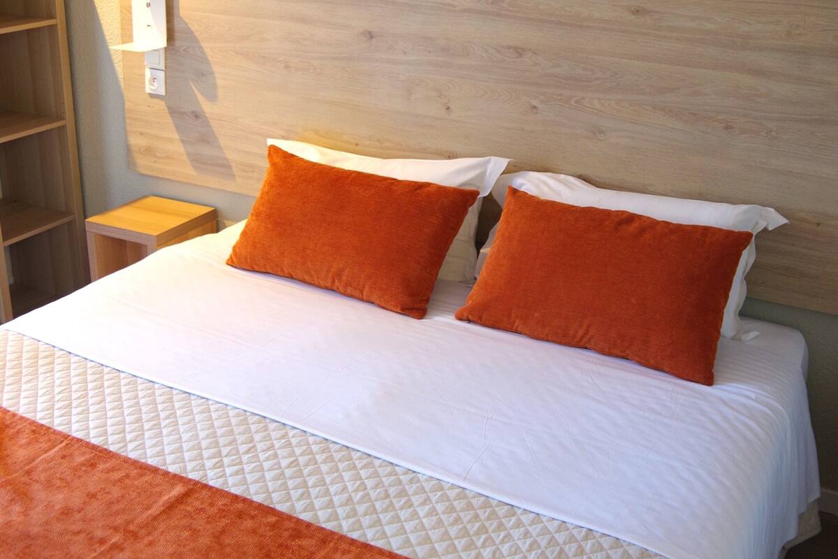 A neatly made bed with two orange pillows and a white duvet, a wooden headboard, and a wall-mounted shelf.