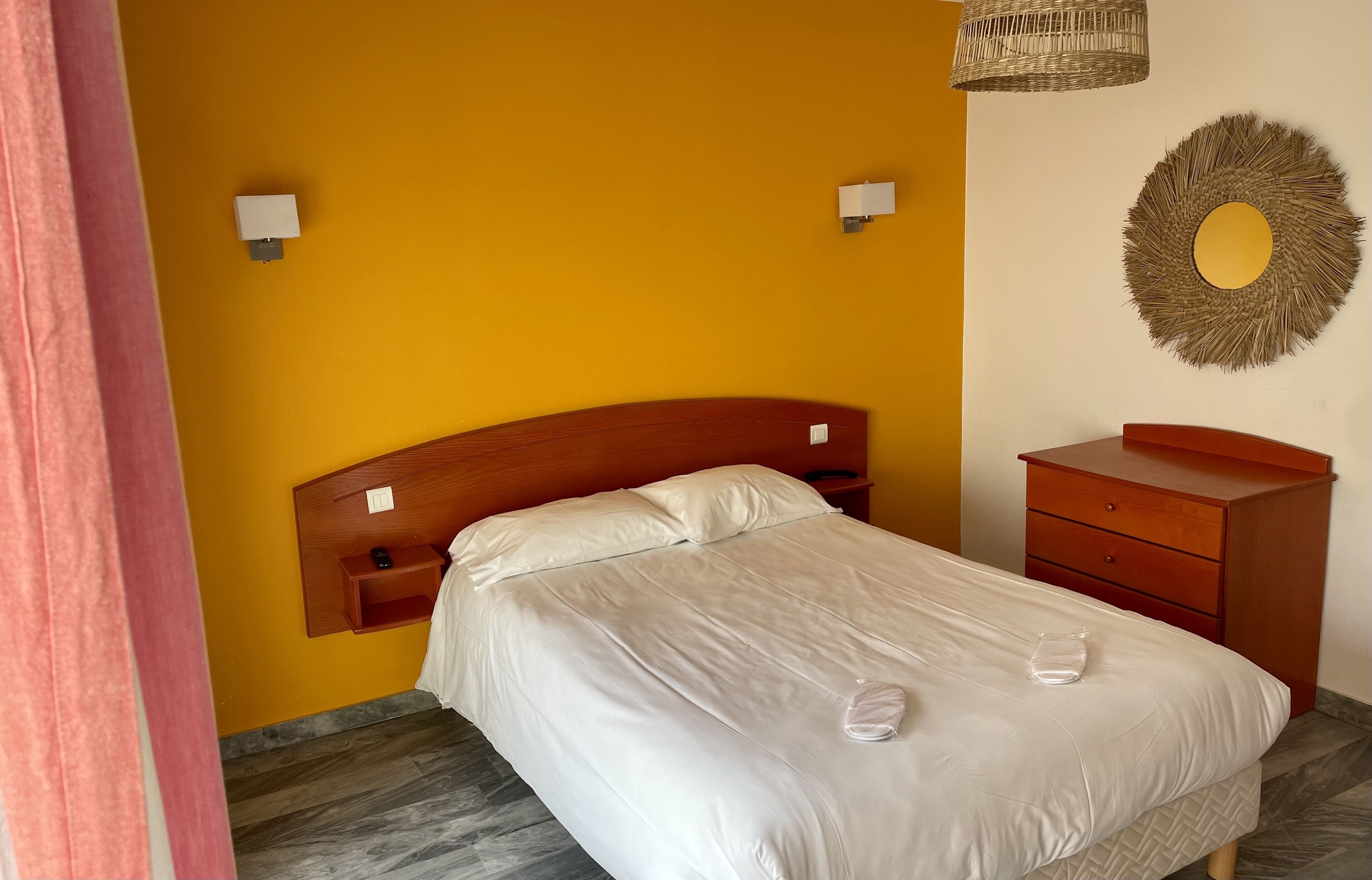 A neatly made bed with white linens, a wooden headboard, a small wooden nightstand with a lamp, a wooden dresser, and a decorative wall mirror.