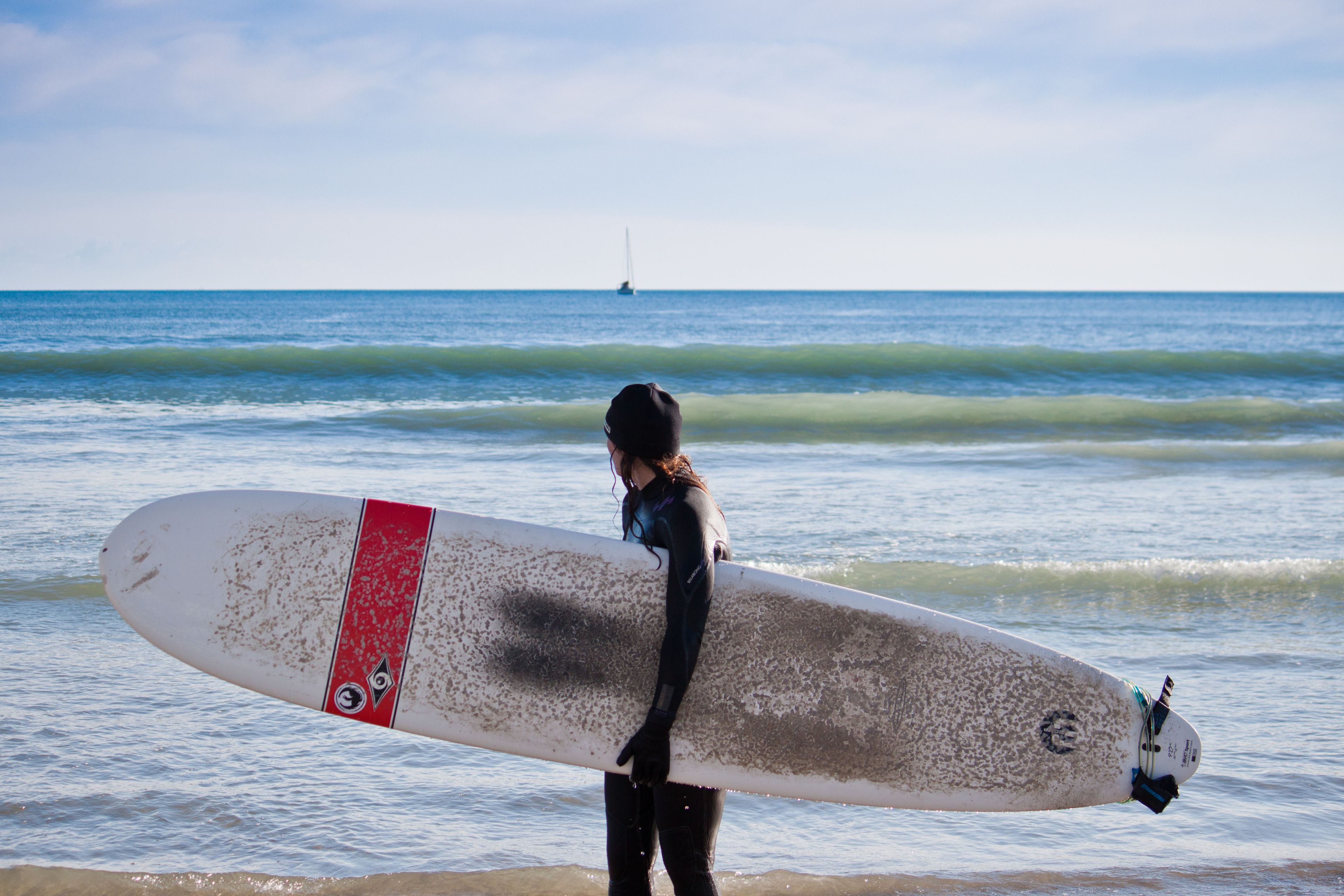 Iemand houdt een surfplank vast op het strand, met golven en een zeilboot in de verte.
