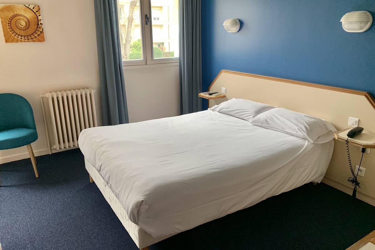 Ein Hotelzimmer mit Bett, Sessel, Nachttisch mit Telefon, Heizkörper und Fenster mit Vorhängen.