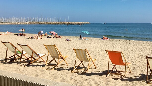 Une plage bordée de rangées de chaises longues et de parasols, une digue rocheuse en arrière-plan et une mer calme.