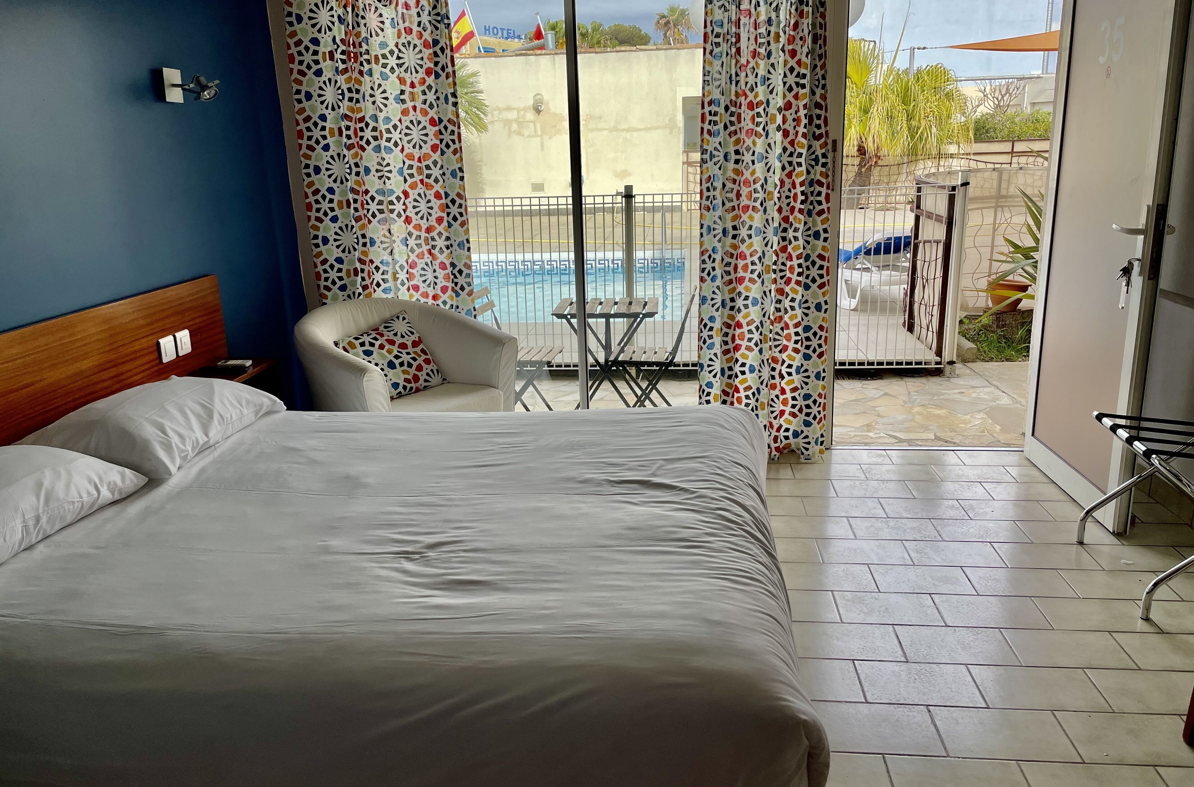 Ein Hotelzimmer mit Bett, Sessel, kleinem Tisch und Blick auf Pool und Außenbereich.