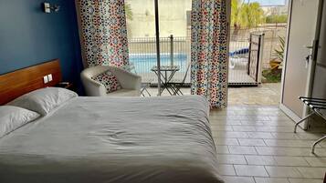 Ein Hotelzimmer mit Bett, Sessel, kleinem Tisch und Blick auf Pool und Außenbereich.