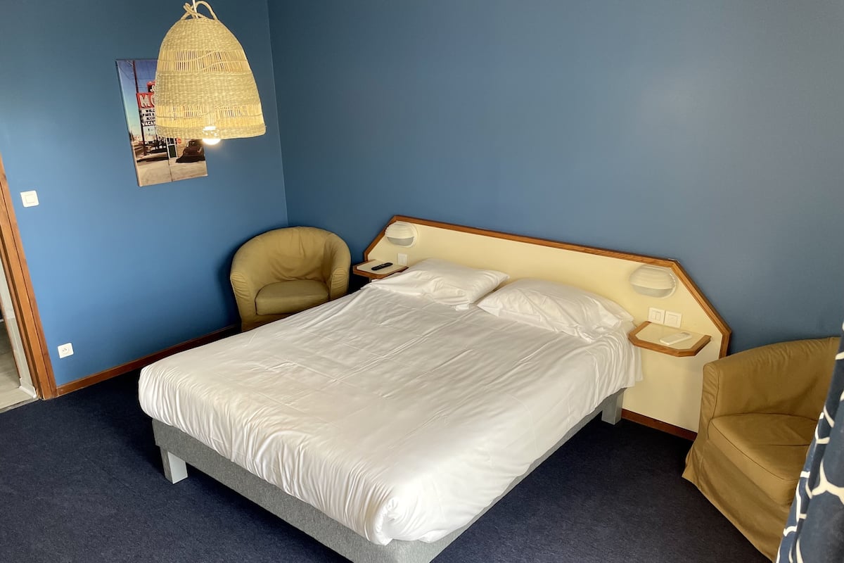 Ein Schlafzimmer mit einem Bett, zwei Stühlen, einem Nachttisch, einer Lampe und einem an der Wand hängenden Bild.