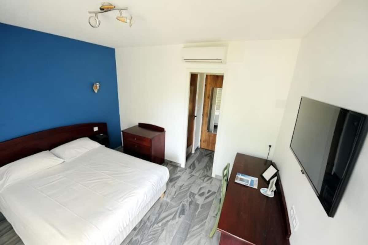 A hotel room with a bed, a desk, a chair, a TV, and an air conditioning unit.