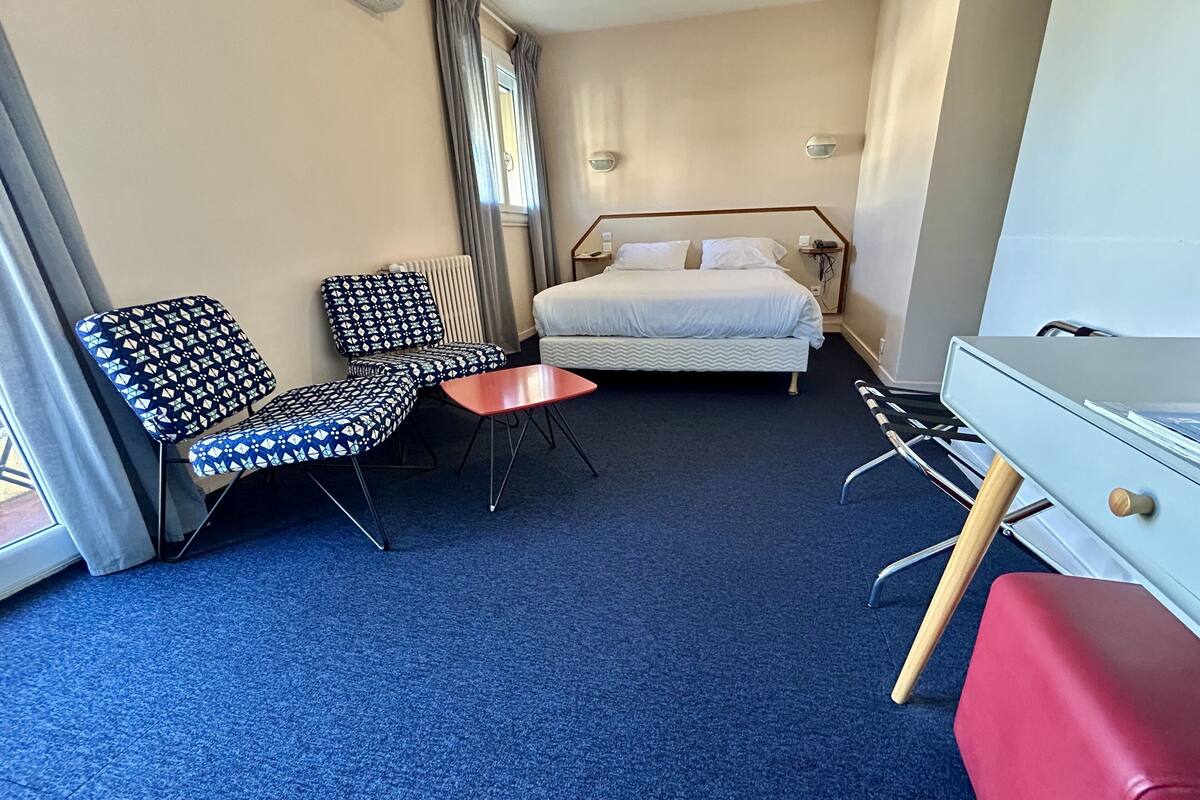Ein Hotelzimmer mit einem Bett, zwei gemusterten Sesseln, einem kleinen Tisch, einem Schreibtisch und einer roten Bank.