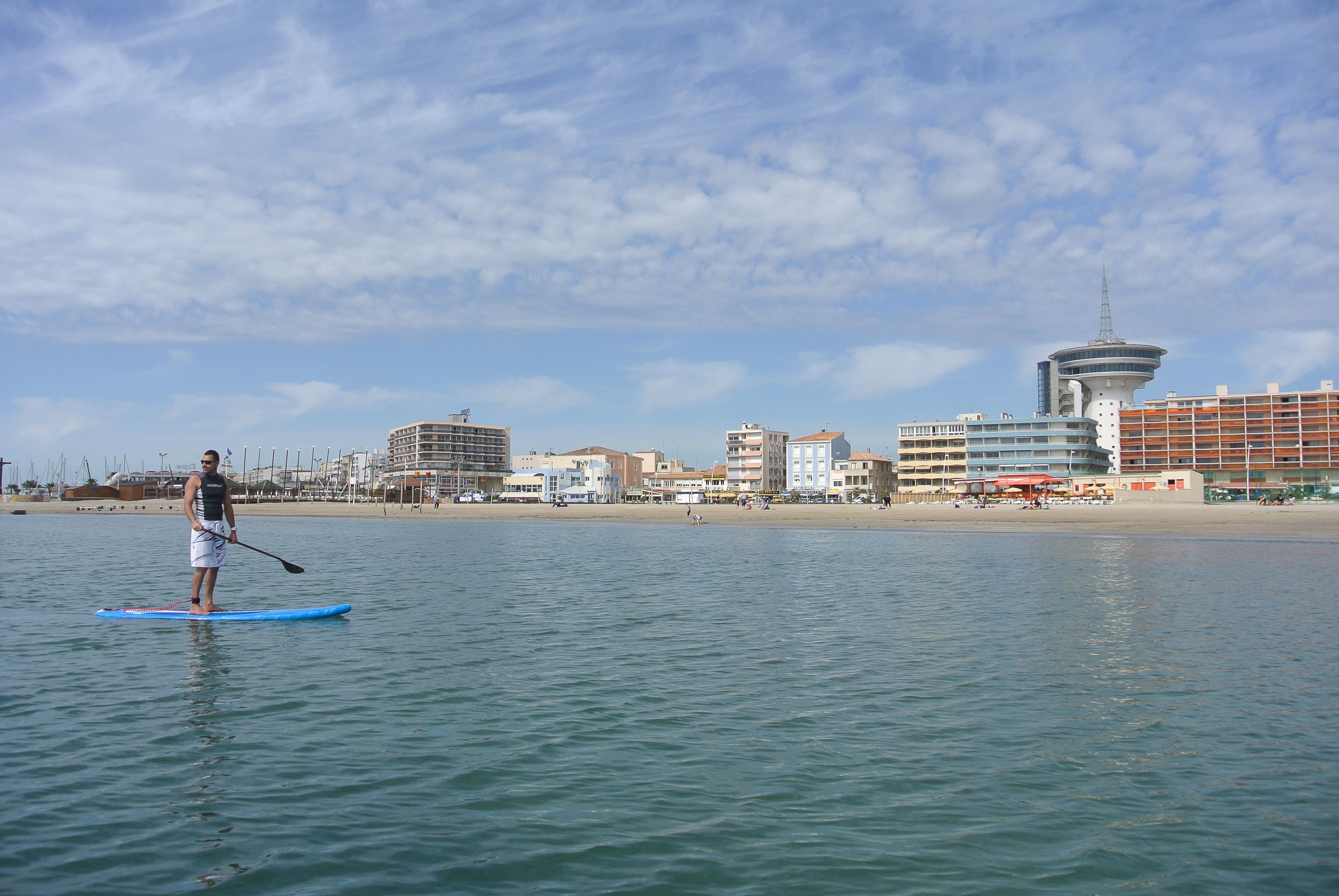 A person paddleboarding on a calm body of water with a cityscape in the background.