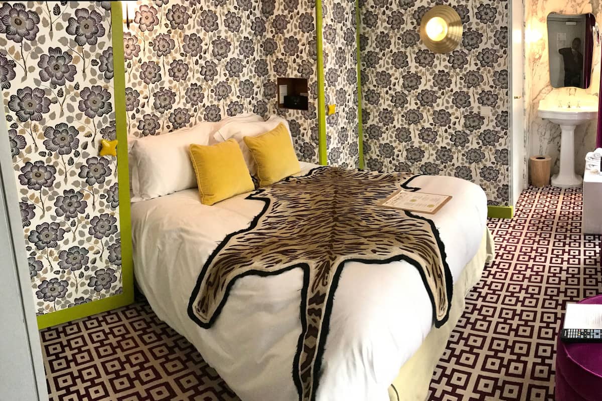 A bedroom with a bed, pillows, a blanket, a bedside table with a book and remote, a mirror, and a bathroom visible through an open door.
