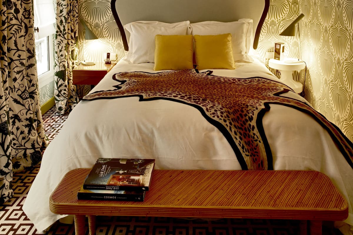 A bedroom with a patterned wallpaper, a bed with white and yellow pillows, a bedside table with a book, and a lamp on the nightstand.