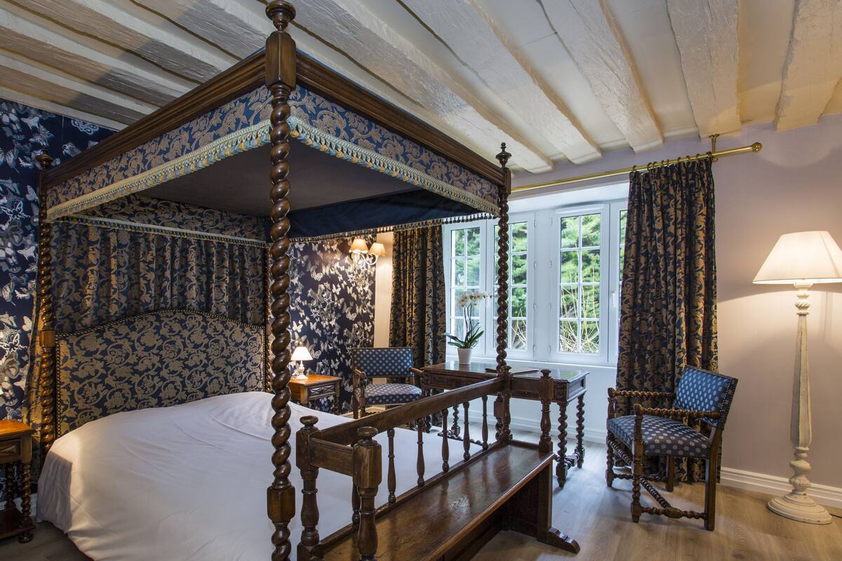 Ein Himmelbett mit floraler Kopfteil, eine hölzerne Bank, ein Stuhl und eine Lampe.