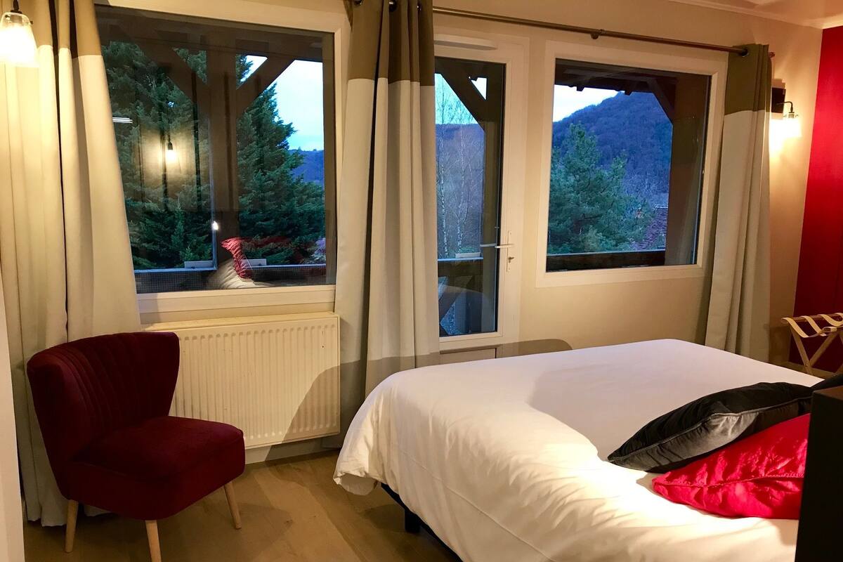 Ein Schlafzimmer mit einem Bett, einem roten Sessel und zwei mit Vorhängen versehenen Fenstern.