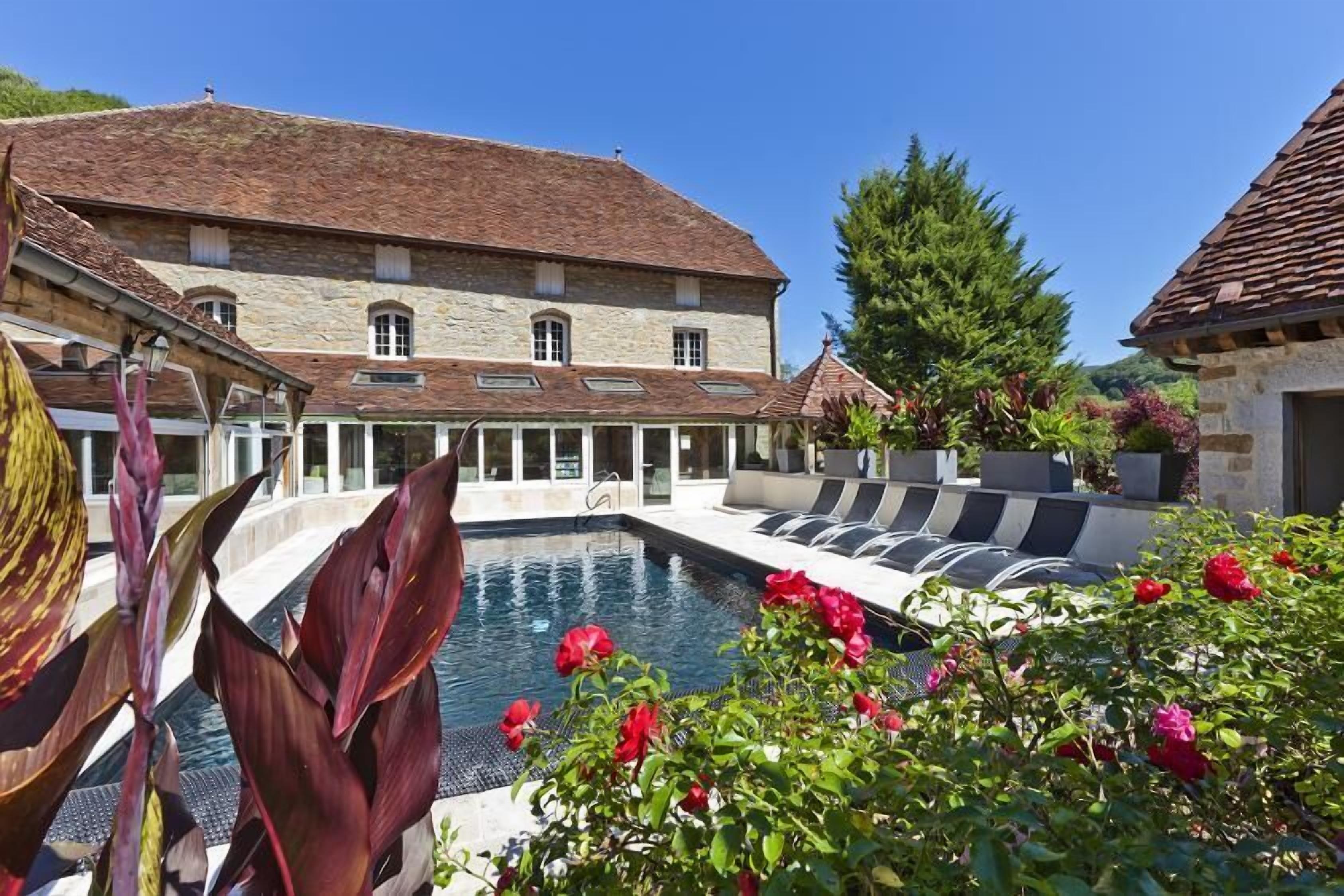 A pool area with a stone building in the background, surrounded by lush greenery and flowers.