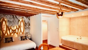 A bedroom with a bed, a wooden ceiling, a chandelier, a bathroom with a bathtub, and a patterned wall. - Castel Damandre (Les Planches-pres-Arbois)