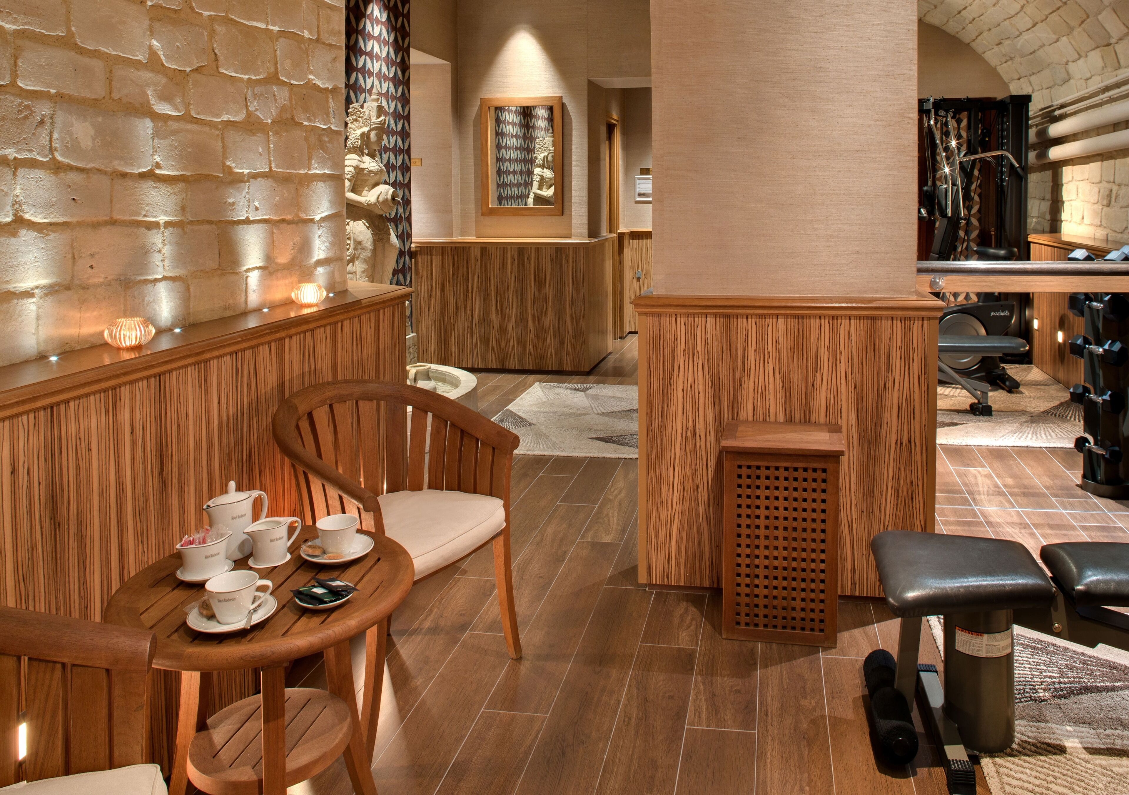 A wooden reception desk with a mirror, a seating area with chairs and a table, and exercise equipment in the background.