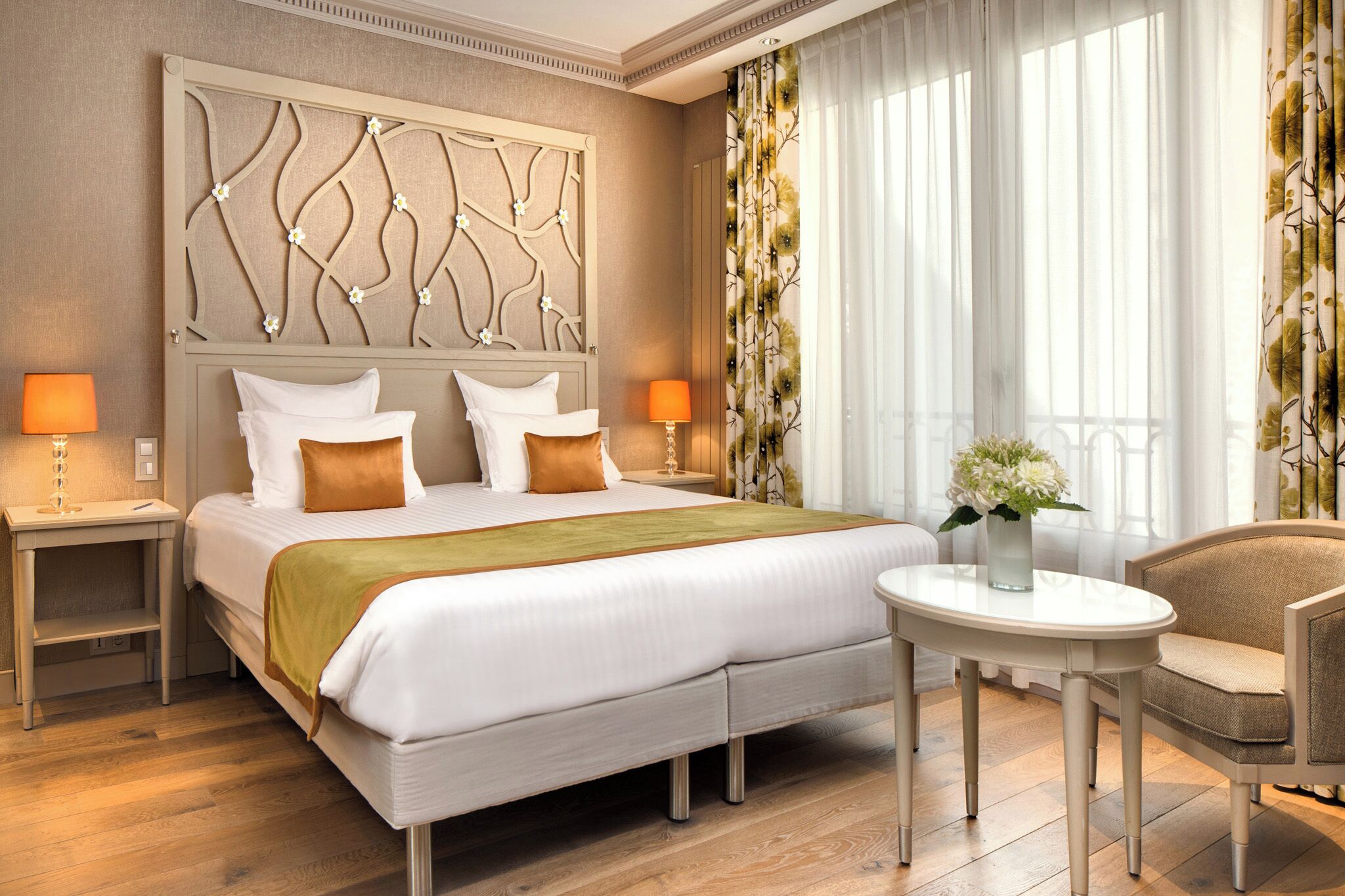 A hotel room with a large bed, two bedside tables with lamps, a round table with a chair, and a decorative headboard.