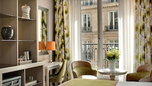 A hotel room with a bed, a desk with a lamp, a chair, a small table with a vase, and a view of a building through a window. - Hotel Rochester Champs Elysées (Paris)