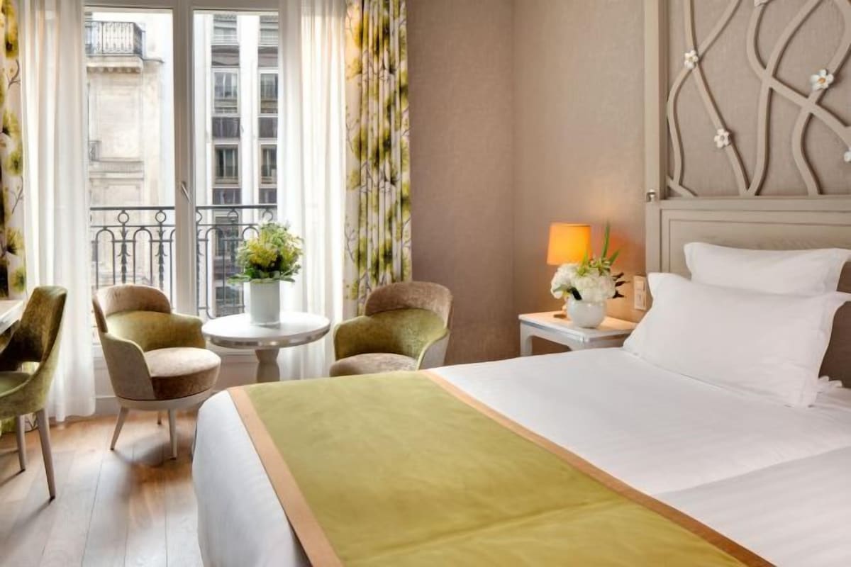 A hotel room with a bed, two armchairs, a small table with a lamp, and a view of the city through a window with curtains.