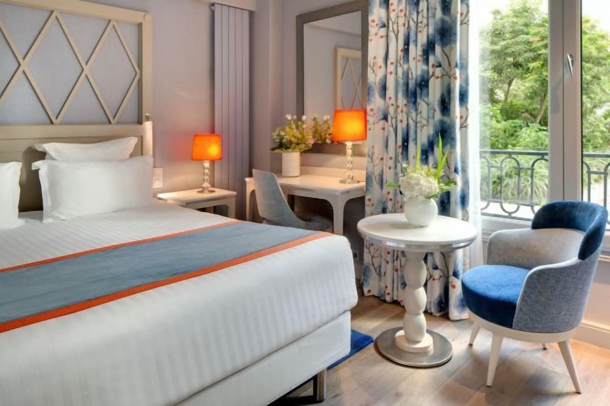 A hotel room with a bed, bedside lamps, a small table with a flower vase, a chair, and a view of greenery through the window.