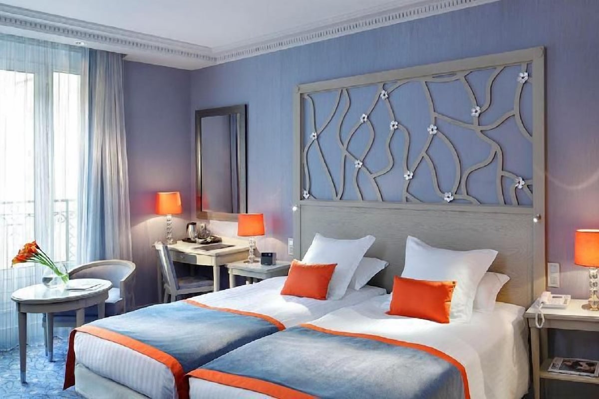 A hotel room with a large bed, two bedside tables, a desk, a chair, and a window with curtains.