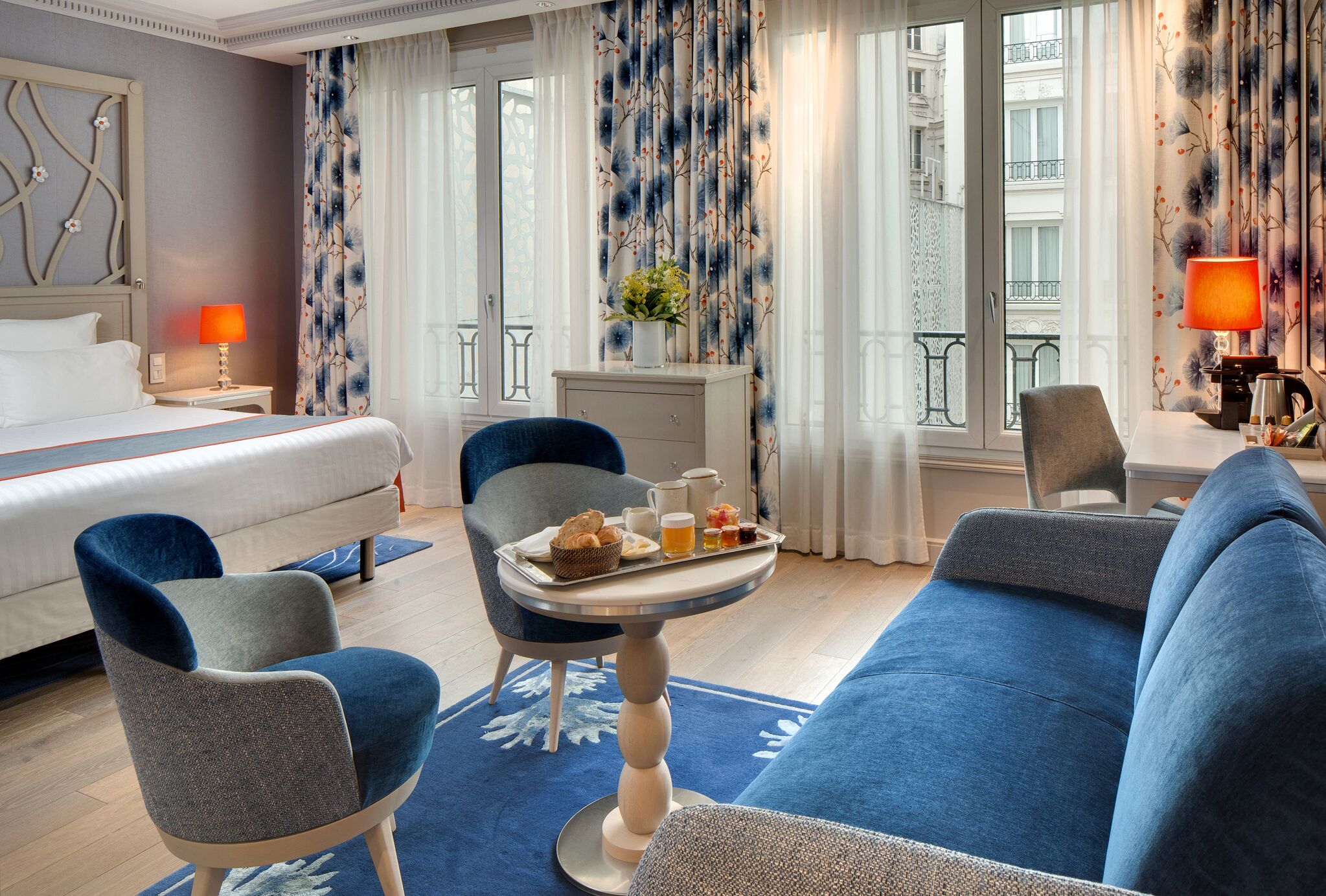 A hotel room with a bed, two armchairs, a sofa, a dining table with breakfast items, and a view of the city through large windows.