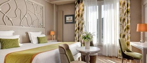 Una habitación de hotel con una cama, dos sillas, una mesa redonda y un ventanal con cortinas.