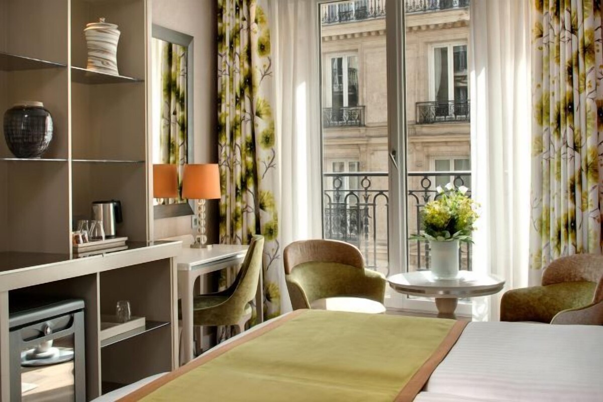 A hotel room with a bed, a desk, a chair, a small table with a vase of flowers, and a view of a building through a window.