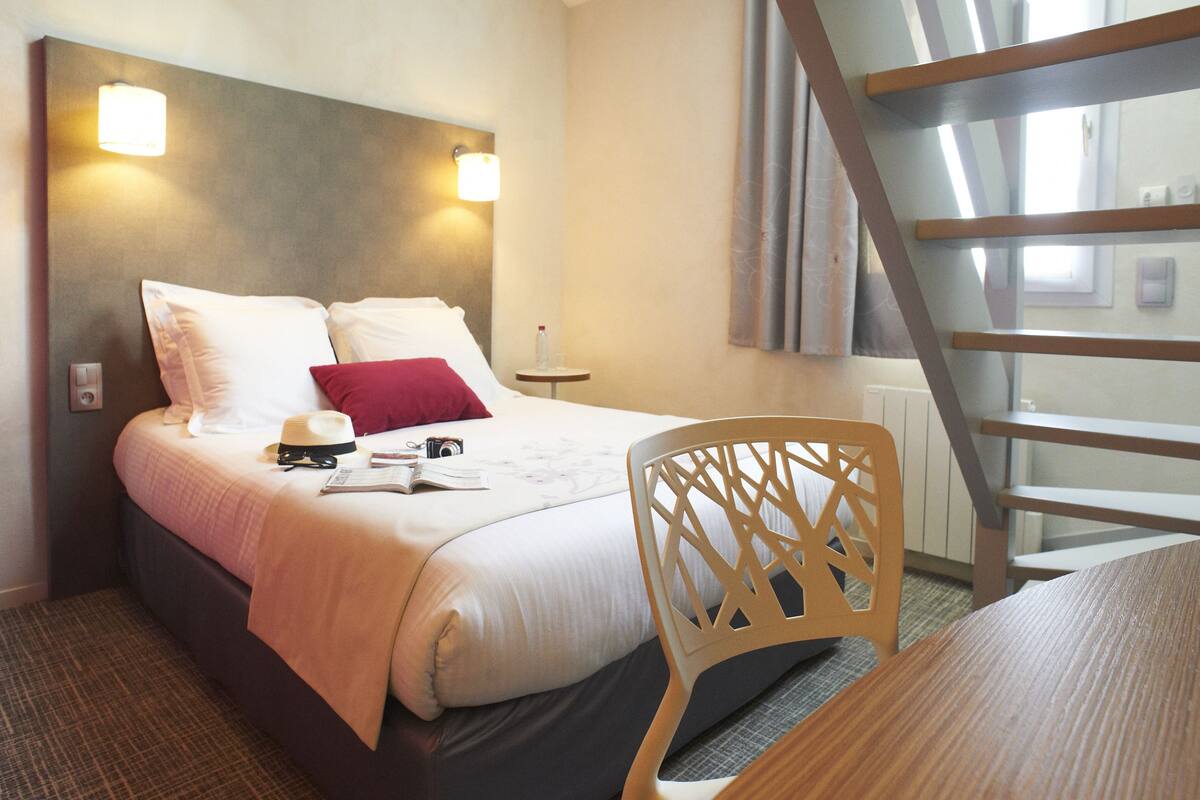 Hotellihuone, jossa on sänky, tuoli, pöytä ja ikkuna, josta on näkymä rakennukseen.