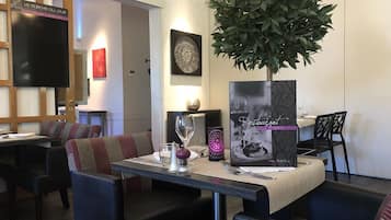 En restauranginteriör med ett bord dukat för två personer, en stor krukväxt och ett menyställ.
