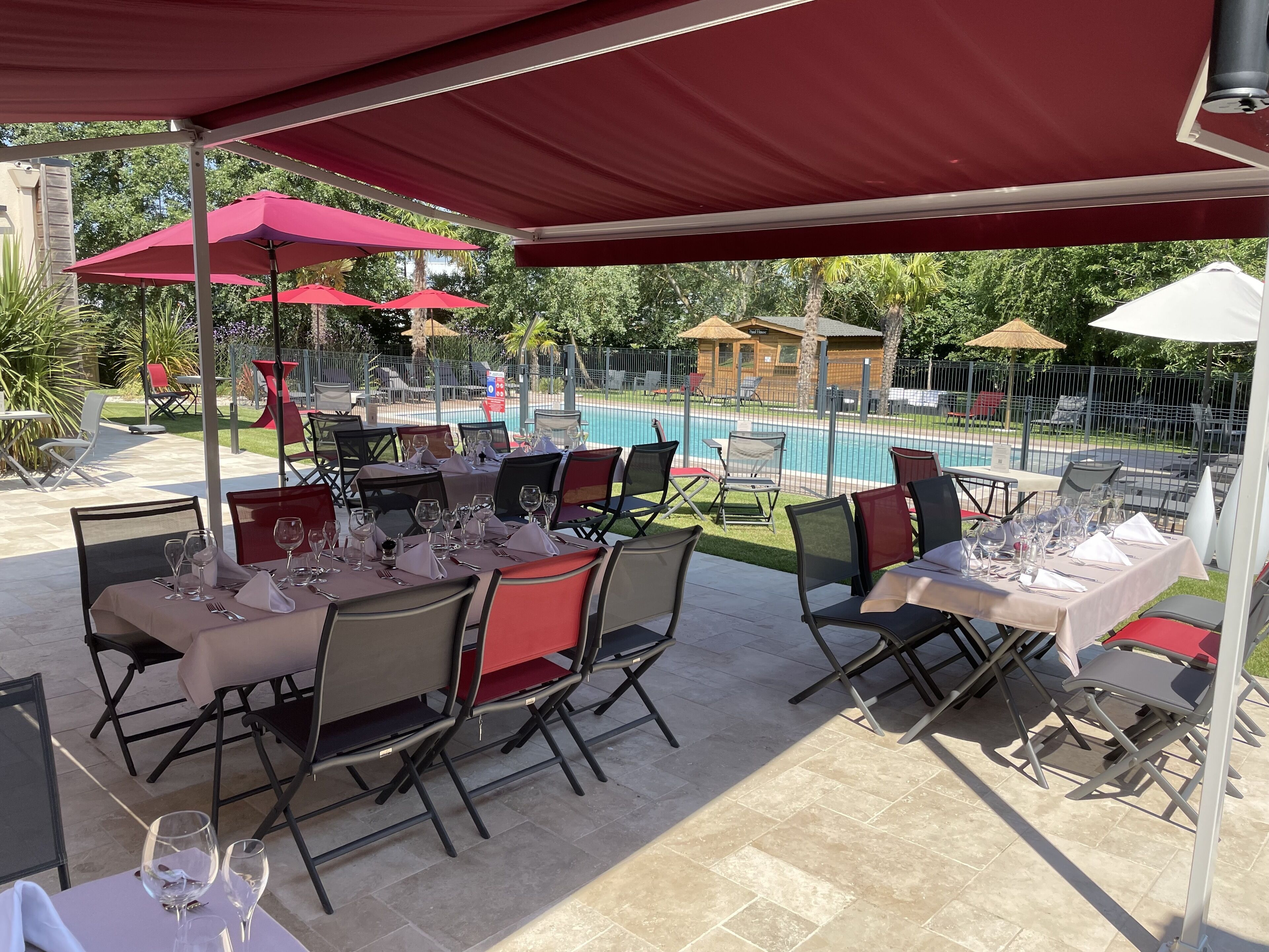 Un área de comedor al aire libre con mesas dispuestas para una comida, rodeada de sombrillas rojas, y una piscina al fondo.