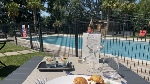 Una mesa de comedor con un plato de comida, una copa de vino y un menú. La mesa está al aire libre, con una piscina al fondo.