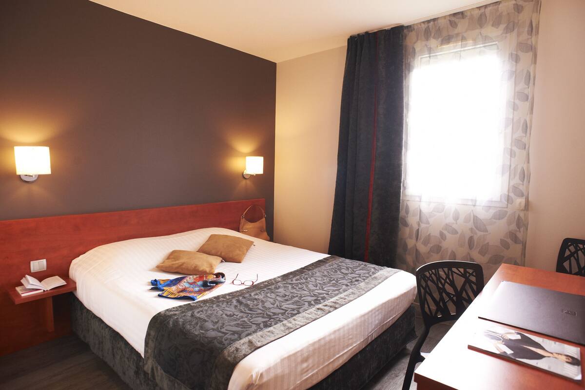 Hotellihuoneessa on sänky, työpöytä ja tuoli. Seinälle on asennettu kaksi valaisinta, ikkunassa on verhot ja sängyllä on käsilaukku.