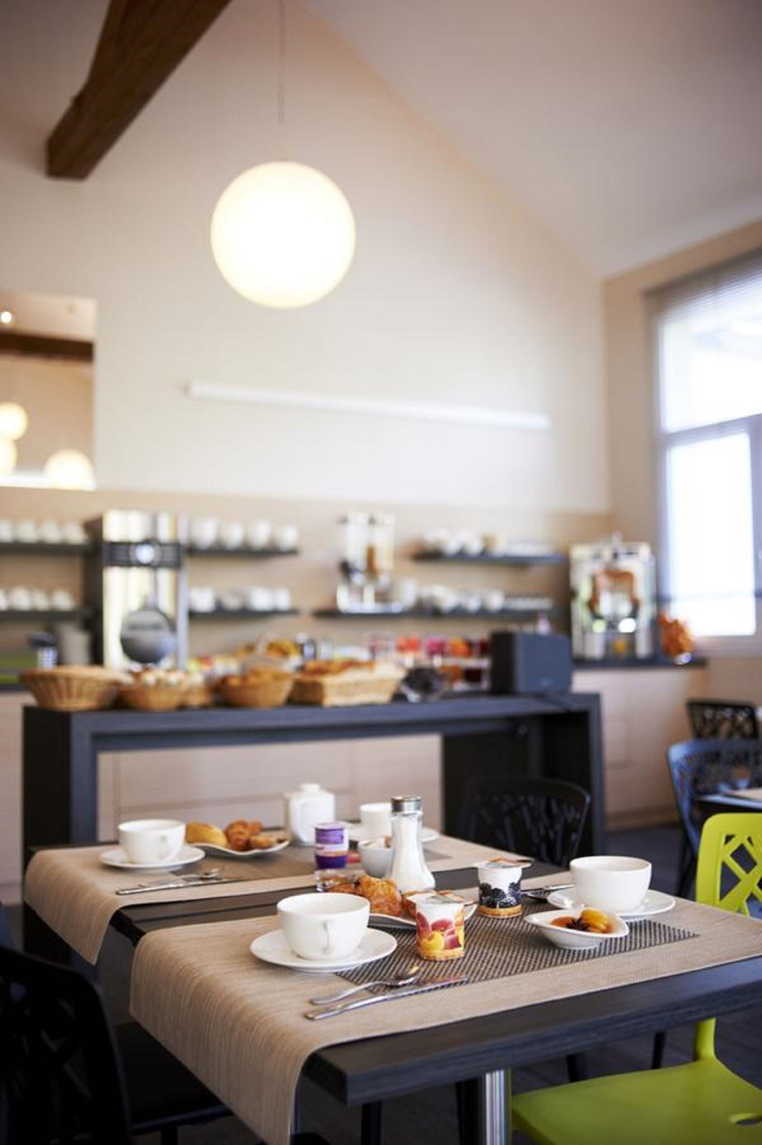 A breakfast setup with a table set for two, a coffee machine, and a variety of pastries and fruits on display in the background.